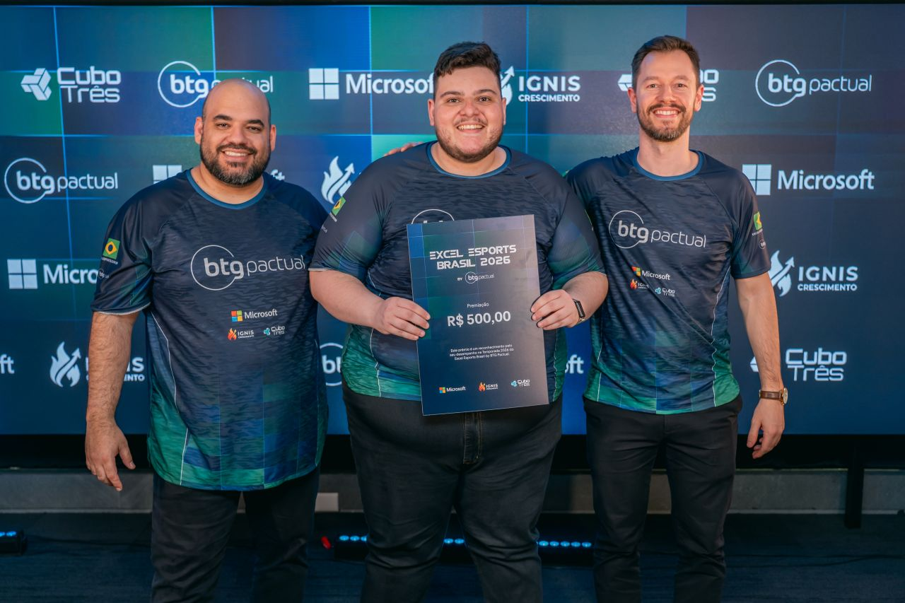 Gabe entre dois participantes, segurando uma placa de premiação do Excel Esports Brasil 2026.